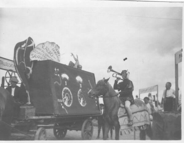 Mustavalkoisessa kuvassa juhlakulkue, jossa hevosen selässä torvea soittava hovimies, hänen takanaan esittelykärryt.